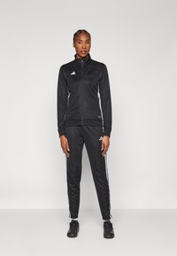 Survêtement de sport noir en tissu lisse ; veste zippée avec bandes latérales et logo ; pantalon assorti avec accents blancs sur les côtés et chevilles resserrées.