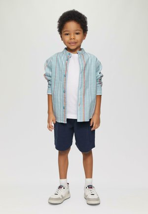 Chemise à boutons rayée bleu clair sur un t-shirt blanc, assortie à des shorts navy et des baskets blanches. Cheveux courts et bouclés.