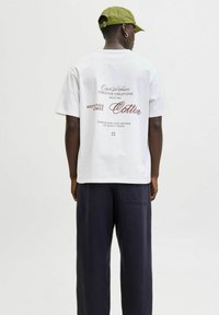 Hvid bomulds t-shirt med trykt tekst på ryggen i bordeaux, kombineret med løstsiddende marineblå bukser. T-shirten har rund halsudskæring og korte ærmer.