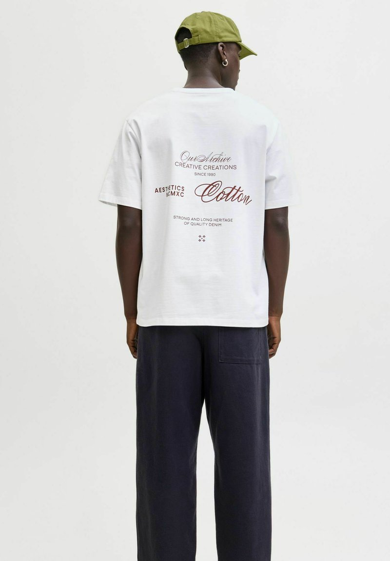 Hvid bomulds t-shirt med trykt tekst på ryggen i bordeaux, kombineret med løstsiddende marineblå bukser. T-shirten har rund halsudskæring og korte ærmer.