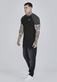 Schwarzes kurzärmeliges T-Shirt mit grauen Raglanärmel, versehen mit einem Logo auf der Brust. Getragen mit dunklen Jeans und weißen Sneakers.