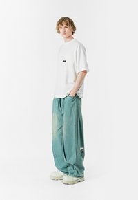 Camiseta blanca de gran tamaño con un bolsillo en el pecho y un logo negro, combinada con pantalones de mezclilla de pierna ancha azul claro y calzado blanco grueso.