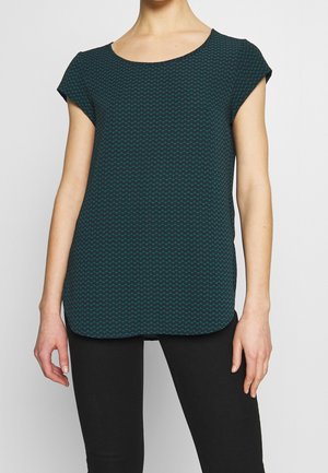 Femme portant un haut à manches courtes vert foncé à motifs avec un col rond, associé à un pantalon noir moulant.