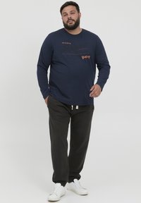Mørkeblå langermet T-skjorte med oransje tekst og prikker, kombinert med svarte bukser og hvite joggesko mot en ensfarget bakgrunn.