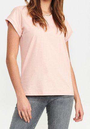 Kobieta w jasnoróżowej koszulce z krótkim rękawem i podwiniętymi mankietami oraz szarych dżinsach, stojąca na jednolitym jasnym tle.