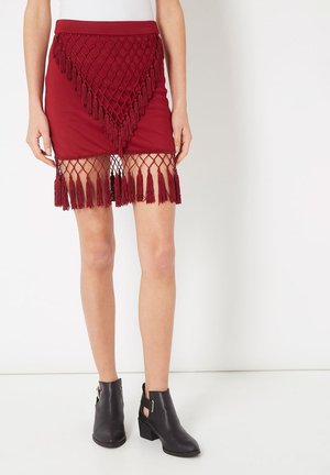 Falda burdeos con flecos de red y borlas en el dobladillo. Presenta una silueta ajustada y una cinturilla elástica. Botines negros completan el conjunto.