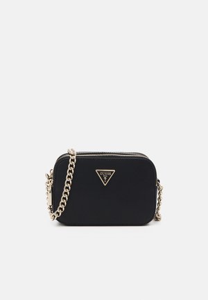 Schwarze Guess Crossbody-Tasche mit goldener Kettenriemen und dreieckiger Logo-Plakette vorne, vor einem einfarbigen hellen Hintergrund.