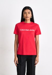 Červené bavlnené tričko s klasickým výstrihom a krátkymi rukávmi, s výrazne potlačeným logom „Calvin Klein Jeans“ v bielej farbe na hrudi.