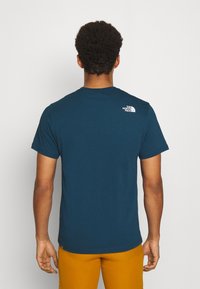 Muž s krátkými kudrnatými vlasy, oblečený v modrém tričku The North Face a hořčicových kalhotách, stojící čelem k obyčejné bílé zdi.