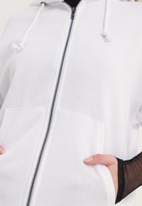 Biela mikina s predným zipsom, šnúrkami a prednými vreckami. Mäkkučký materiál, voľný strih, s čiernym detailom zipsu.