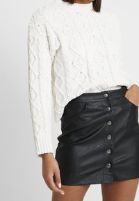 Pull en maille blanche à col haut, associé à une mini-jupe noire en simili cuir avec des boutons-pression à l'avant et des poches.