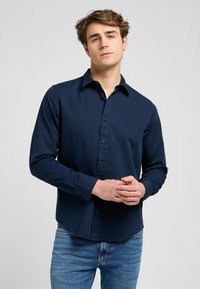 Meripanjang berwarna biru navy terbuat dari katun, dilengkapi dengan kerah kancing, kancing depan, dan bordir logo yang halus di dada.