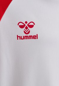 Hvid sportstrøje med røde accenter på skuldrene og et rødt "hummel"-logo med et karakteristisk symmetrisk design.