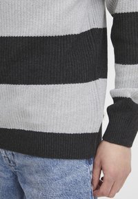 Sweter dzianinowy z poziomymi czarnymi, szarymi i białymi paskami. Ściągacze na mankietach i na dole, wykonany z teksturowanej tkaniny, noszony z dżinsami.