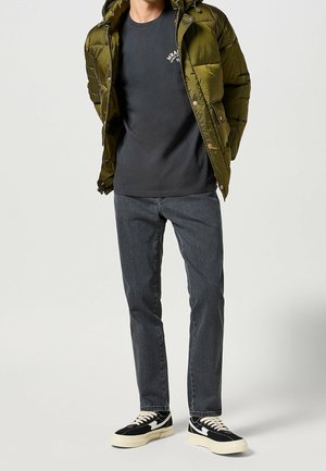 Hombre con chaqueta acolchada verde oliva sobre camiseta negra con estampado, jeans gris oscuro y zapatillas negras y blancas, de pie contra un fondo claro.