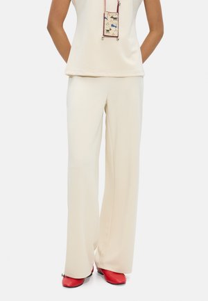 Kobieta ubrana w kremowe spodnie o szerokich nogawkach, pasujący bezrękawnik, czerwone buty z kwadratowym noskiem oraz małą torebkę w zwierzęce wzory na sznurku.