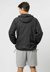 Mees, kes on seljaga vaataja poole, kannab musta kapuutsiga jopet, millel on kiri "U.S. Polo Assn." ja helehallid lühikesed püksid, taust on ühevärviline.