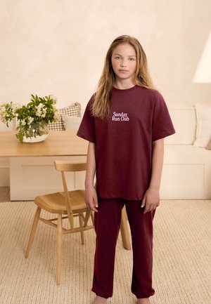 Jeune fille debout à l'intérieur portant un t-shirt et un pantalon bordeaux assortis "Sunday Run Club", avec une chaise et une table en bois en arrière-plan.