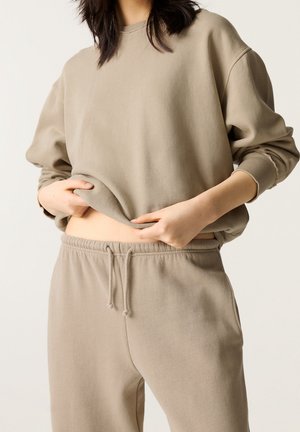 Mujer con sudadera beige holgada y pantalones deportivos a juego, levantando ligeramente la camiseta para mostrar el abdomen, con fondo claro y liso.