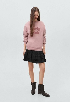 Mistige roze sweatshirt met vetgedrukte tekst, zwarte gelaagde rok met kant, en bruine enkellaarzen. Eenvoudige, casual outfit met contrasterende kleuren.
