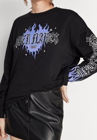 Svart långärmad tröja med en flamediagram i lila och vit text. Designen inkluderar ytterligare text i en intrikat stil på ärmen.