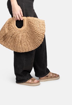Personne portant un pantalon noir et des sandales beiges, tenant un grand sac à main en paille tissée avec une anse rectangulaire.