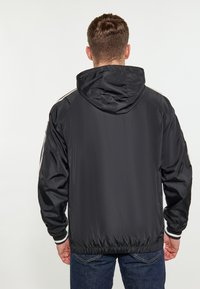 Giacca leggera nera con cappuccio, polsini e orlo elasticizzati. Presenta due righe laterali sulle maniche e una texture liscia e lucida.