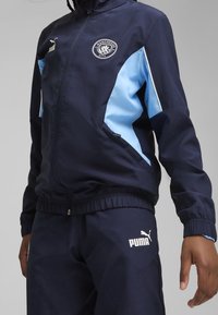 Veste de course bleu marine avec des accents bleu clair, logo PUMA et emblème de Manchester City. Manches et taille élastiques ; texture de tissu lisse.