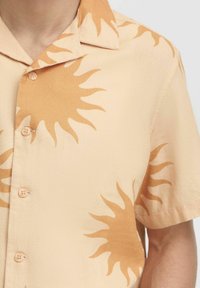 Kurzarmhemd aus hellem beige Stoff mit strukturierter Oberfläche, mit einem Sonnenmuster in dunklerem Beige. Knopfverschluss vorne.