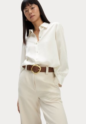 Witte satijnen shirt met lange mouwen en knoopmanchetten, gecombineerd met hoogwaardige beige broeken en eenbruine riem met een grote gouden gesp.