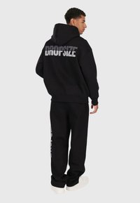 Homme en hoodie noir et pantalon avec le texte "DROPSIZE" dans le dos et sur la jambe, portant des baskets blanches, se tenant devant un fond clair.