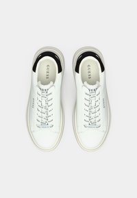 Fehér alacsony szárú sneaker, fekete sarokrész-kiegészítőkkel és "GUESS" feliratos fűzőkkel, felülről nézve, fehér háttér előtt.