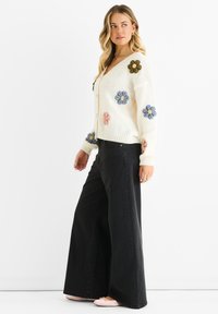 Knitted cream cardigan with floral embroidery in blue, pink, and green. Paired with high-waisted black wide-leg jeans and light-colored footwear.
