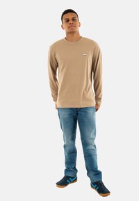 Camicia beige in cotone a maniche lunghe con scollatura rotonda e piccolo dettaglio grafico, abbinata a jeans in denim azzurri e sneaker nere con dettagli blu.