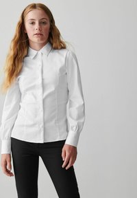 Camicetta bianca a maniche lunghe con colletto, presenta una vestibilità aderente, dettagli sulle cuciture frontali sottili e polsini con bottone. Abbinate a pantaloni neri.