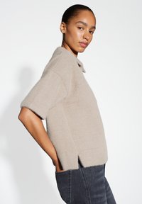 Pull en tricot beige à manches courtes, avec un col et une coupe ample. Son texture douce et ses fentes latérales sont associées à un jean foncé.