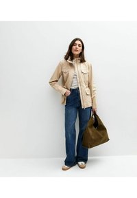 Beige crop jack met twee voorzakken, gestreept shirt, blauwe flared jeans en een groen suède handtas. Bruine en witte schoenen maken de look compleet.
