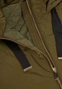 Intérieur d'une veste matelassée vert olive, avec un motif de piquage, une fermeture éclair dorée, et des accents en tissu noir sur les bords. Texture lisse.