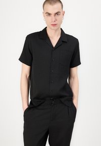 Chemise noire à manches courtes avec une poche, présentant un subtil motif hexagonal, un devant boutonné et une coupe décontractée, assortie à un pantalon correspondant.