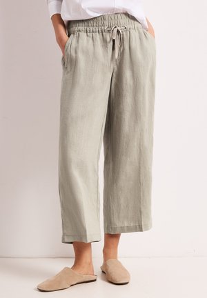 Beige linnedragt med vide ben og elastisk snøre i taljen, båret med beige slip-on sko og hvid indstikket skjorte. Hænderne i lommerne.