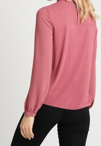 Rosa blus med långa, puffade ärmar, rund halsringning och en subtil volangdetalj vid kragen. Slätt, lätt tyg.