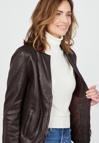 Lächelnde Frau mit schulterlangem Haar, die einen weißen Rollkragenpullover und eine dunkelbraune, über ihre blauen Jeans offene Lederjacke trägt.