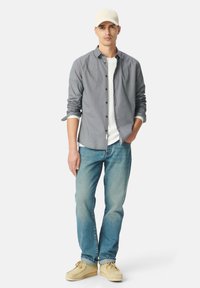 Camicia grigia a maniche lunghe con un motivo sottile, t-shirt bianca sotto, jeans azzurri chiari e scarpe beige. Un cappellino bianco completa l'outfit.