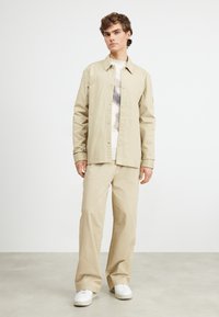 Chaqueta de camisa beige con corte recto, que presenta botones a presión y un suéter ligero debajo, combinada con pantalones holgados beige y zapatillas blancas.