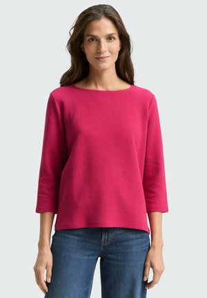 Donna con i capelli castani lunghi che indossa un top rosa brillante a maniche tre quarti e dei jeans bleu, in piedi davanti a uno sfondo chiaro e semplice.