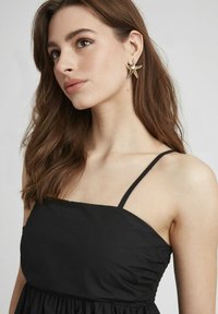 Robe noire à fines bretelles, présentant un corsage froncé et un bas fluide. Une boucle d'oreille en forme d'étoile de mer dorée ajoute une touche de détail à ce style.