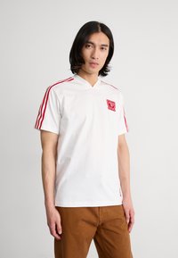 Camisa polo branca de manga curta com decote em V, apresentando detalhes em vermelho com três riscas nos ombros e um logo vermelho no lado esquerdo do peito.