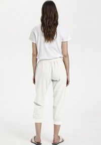 Kvinna med långt, vågigt brunt hår står vänd bort, iförd en vit kortärmad skjorta, vita cropped-byxor och beige slidesandaler.