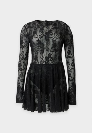 Femme portant une robe longue en dentelle noire à manches longues et des bottines noires, posant devant un fond clair uni.