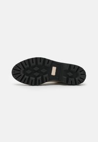 Suela de goma negra de un zapato con surcos profundos, patrones en forma de diamante y una pequeña inserción rectangular beige, mostrada sobre un fondo blanco.